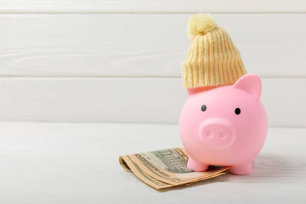 Piggy bank with money.Piggy bank with dollars and winter hat on white texture wood.Heat saving concept. The concept of saving heating.Banking and savings. Place for text, space for copy.