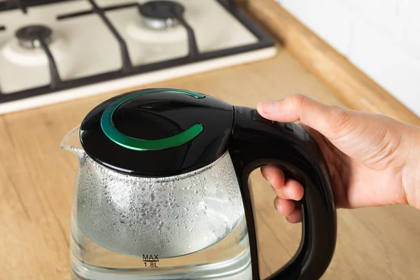 Close-up of a woman pressing the power switch on an electric kettle.Save energy at home.A modern electric transparent kettle on a wooden table in the kitchen. A kettle for boiling water and making tea.