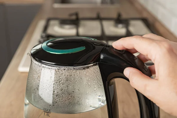 Close-up of a woman pressing the power switch on an electric kettle.Save energy at home.A modern electric transparent kettle on a wooden table in the kitchen. A kettle for boiling water and making tea.
