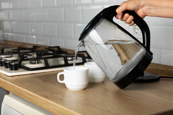 A man makes tea using boiling water from an electric kettle in the kitchen at home. It's time to have breakfast and drink tea. Modern electric kettle on a wooden table. Kettle for boiling water.