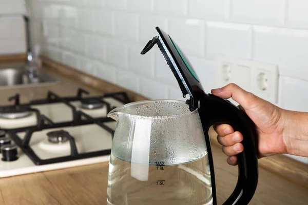A hand opens the lid of an electric kettle. A modern electric transparent kettle on a wooden table in the kitchen. A kettle for boiling water and making tea. Home appliances for making hot drinks.