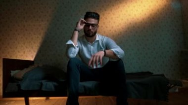 Genç adam başı eğik ve üzgün bir şekilde yatakta oturuyor.