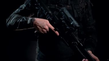 Karanlık bir geçmişi olan, kamuflajlı bir asker taktik tüfeğini kemerine takıyor.