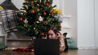 Genç bir kız, yılbaşı akşamı süslü bir Noel ağacının altında dizüstü bilgisayarda bağımsız olarak çalışıyor.