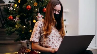 Solunum maskeli tatlı bir kız Noel arifesinde dizüstü bilgisayarda çelenklerle süslenmiş bir Noel ağacının arka planına karşı çalışıyor.