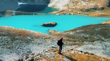 Sırt çantalı gezgin bir tepede duruyor ve dağdaki çayırlara ve güzel bir dağ gölüne hayran. İnsansız hava aracı görüntüleri. Paralax videosu. Dolly yakınlaştır.