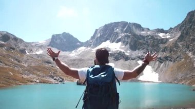 Sakallı bir dağcı dağın tepesinde bir dağ gölünün yanında duruyor ve zafer kazanıyor.