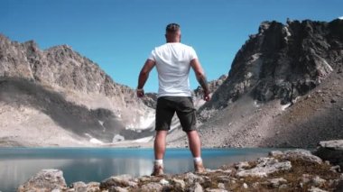 Sakallı ve atletik vücutlu bir erkek dağın tepesinde bir dağ gölünün kenarında duruyor ve kollarını destansı bir şekilde açıyor.