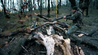 Sakallı ve silahlı bir adam ormanda büyük bir ağacın üzerinden atlıyor. Yavaş çekim