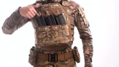 Çoklu ambalajlı kamuflajlı özel harekat subayı, plaka taşıyıcısındaki dolu bir şarjörden beyaz arka planı olan bir şarjörü çıkarıyor.