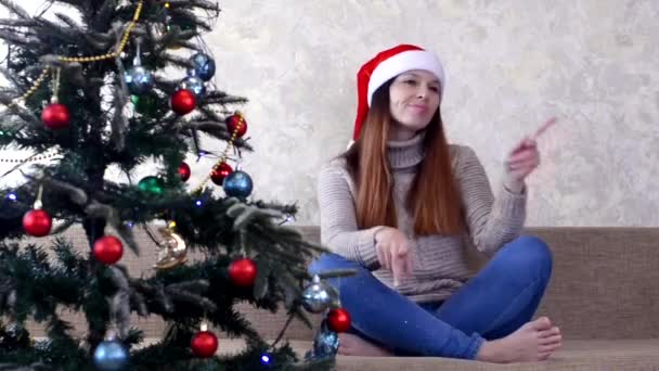 Kaukasische Millennial junge Frau mit Weihnachtsmann Hut feiert Weihnachten zu Hause, Weihnachtsbaum dekoriert rote Spielzeugkugeln und Lichtergirlanden drinnen — Stockvideo