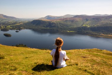 Beautiful girl in a light dress with long hair sits on the edge of a mountain overlooking a lake, summer England, Lake District