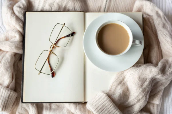 Open brown book with blank pages and mug of cocoa with saucer on warm beige sweater, square glasses. Autumn mood, hygge and comfort