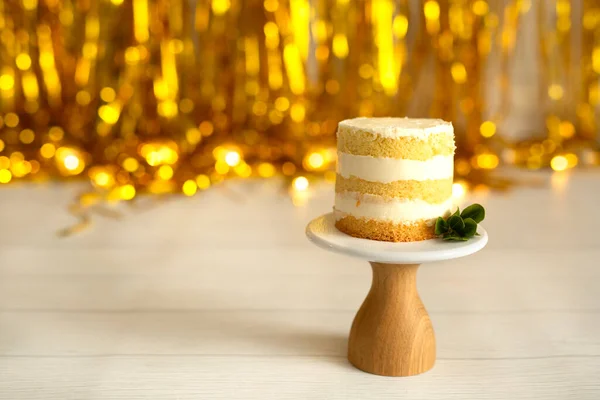 Celebration. A small cake, bare cakes of dough and white cream, on a brown wooden stand on a blurred background of gold decor. Selective focus