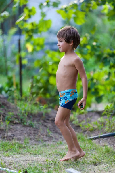 Crianças atividade ao ar livre. Sorria menino criança vestindo um azul calções de natação correndo e brincando com respingos de água se divertindo em um quintal em um dia de verão quente ensolarado. Caucasiano menino joga esconder e procurar — Fotografia de Stock