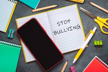 Kabadayılığı bırak. Not defterinin üst görünümü ile metin zorbalığı durdurun, okul malzemeleri ve gri arkaplan üzerinde akıllı telefon