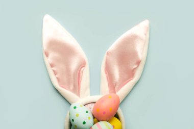 Mutlu Paskalyalar. Renkli paskalya yumurtası beyaz tavşan kulaklarına mavi arka planda boyanmış.