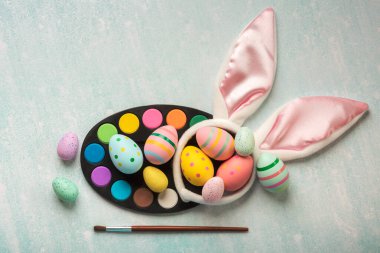 Mutlu Paskalyalar. Renkli paskalya yumurtaları suluboya, fırça ve tavşan kulakları mavi bir masada.