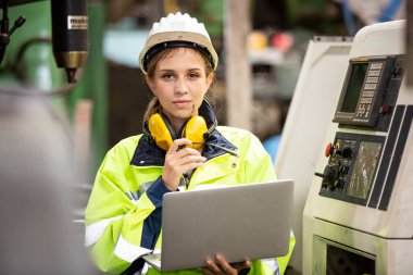 Koruyucu kasklı kadın işçi fabrikada dizüstü bilgisayarla çalışıyor.