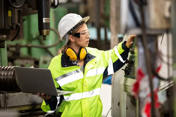 Koruyucu kasklı kadın işçi fabrikada dizüstü bilgisayarla çalışıyor.