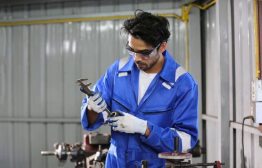 Mavi tulumlu, şapkalı ve eldivenli bir erkek araba operatörü beyaz arabanın kaputunda çalışıyor ve tamirhanedeki motorun çalışabilirliğini kontrol ediyor. Araba bakımı kavramı