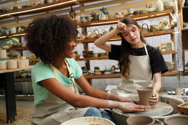 Konsantre olmuş, önlüklü, çömlek direksiyonunda oturan ve ıslak kil kabı şekillendirirken el işi aletleri kullanan güzel bir zanaatkar.