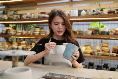 Konsantre olmuş, önlüklü, çömlek direksiyonunda oturan ve ıslak kil kabı şekillendirirken el işi aletleri kullanan güzel bir zanaatkar.