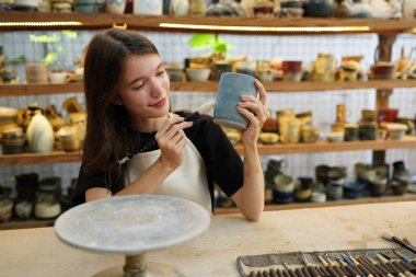 Konsantre olmuş, önlüklü, çömlek direksiyonunda oturan ve ıslak kil kabı şekillendirirken el işi aletleri kullanan güzel bir zanaatkar.