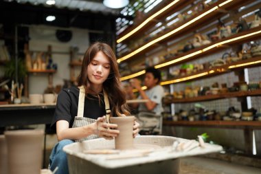 Konsantre olmuş, önlüklü, çömlek direksiyonunda oturan ve ıslak kil kabı şekillendirirken el işi aletleri kullanan güzel bir zanaatkar.