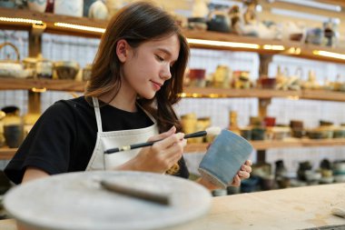 Konsantre olmuş, önlüklü, çömlek direksiyonunda oturan ve ıslak kil kabı şekillendirirken el işi aletleri kullanan güzel bir zanaatkar.