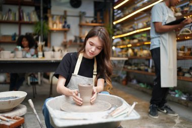 Konsantre olmuş, önlüklü, çömlek direksiyonunda oturan ve ıslak kil kabı şekillendirirken el işi aletleri kullanan güzel bir zanaatkar.