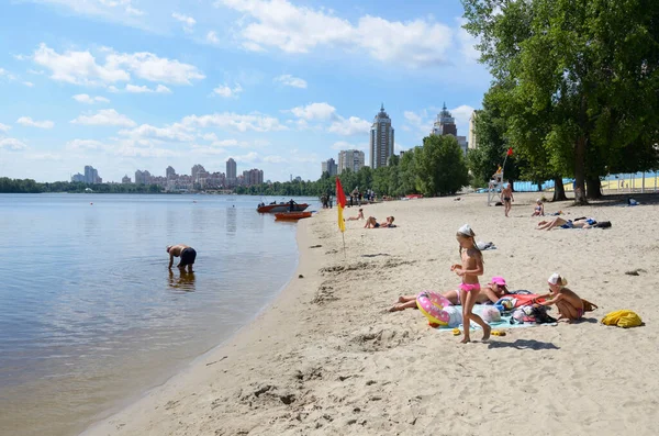 İnsanlar Dinyeper nehrinin sahilinde güneşleniyor, arka planda binalar var.