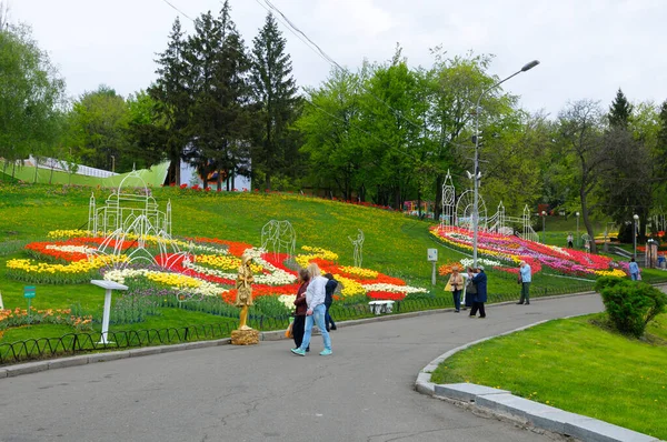 Şehir parkında yürüyen ve çimlere dikilmiş parlak lalelere bakan insanlar. Dünyanın dört bir yanındaki laleler festivalinin açılışı, Şarkı Söyleyen Saha Parkı. 27 Nisan 2018. Kiev, Ukrayna
