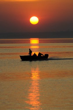 Durgun denizde kızıl gün batımı, üç adamla balığa çıkan bir teknenin siyah silueti.