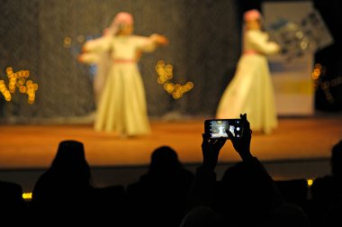 Konser sırasında sahnede dans eden bulanık dansçılar, ellerinde cep telefonlarıyla onları videoya çekiyorlar..