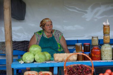 Kırsal gıda pazarı. Ön planda turşulu ve sebzeli konserve kutularıyla tezgahta oturan orta yaşlı kadın sokak satıcısı. 10 Temmuz 2019. Klavdiivka, Kyiv oblast, Ukrayna