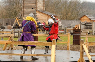 Men reenactors in metal armor of an Old Rus knights reconstructing sword fight, wooden fortress on a background. Kyiv Rus park, Ukraine