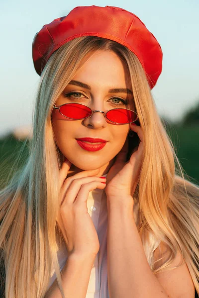 Açık havada mutlu, çarpıcı sarışın kızın moda portresi. Güzel gülümseme. Şık siyah palto, kırmızı bere ve kırmızı gözlük takıyor..