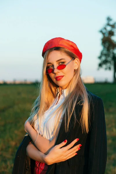 Açık havada mutlu, çarpıcı sarışın kızın moda portresi. Güzel gülümseme. Şık siyah palto, kırmızı bere ve kırmızı gözlük takıyor..