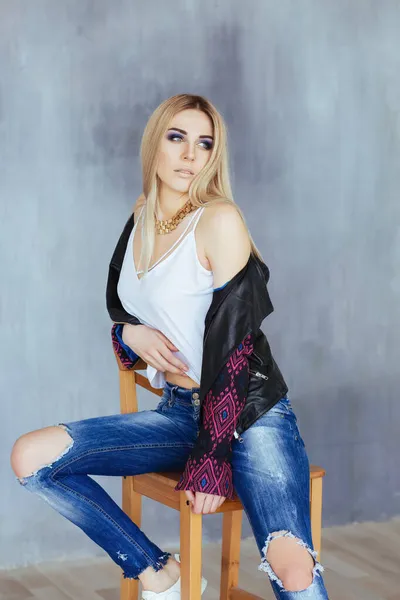 Mladá krásná žena s živým make-up v bílé horní černé bundě a roztržené modré džíny pózovat na židli — Stock fotografie