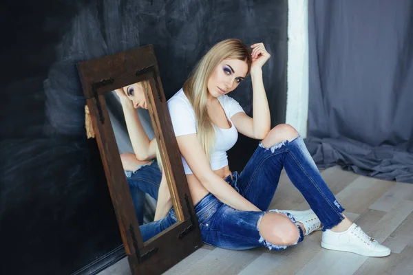 年轻的金发女郎，头戴白色上衣，穿着撕破的蓝色牛仔裤，在镜子前摆姿势 — 图库照片