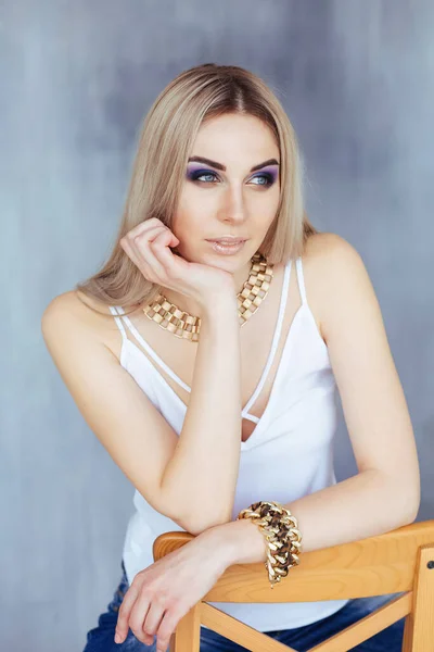 Mladá krásná blondýna žena s živým make-up v bílém top pózování na židli v blízkosti světlé stěny — Stock fotografie