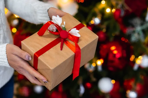 Γυναικεία χέρια με κουτί δώρου κοντά στο χριστουγεννιάτικο δέντρο, κοντινό πλάνο. Παρόντες στα χέρια στη διακόσμηση του νέου έτους Εικόνα Αρχείου