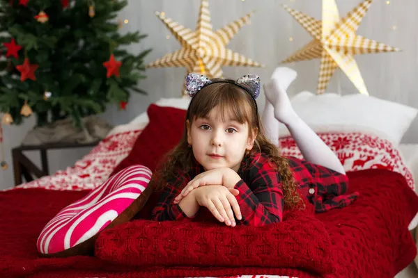Χαριτωμένο κορίτσι παιδί με φόρεμα στο κρεβάτι με χριστουγεννιάτικες διακοσμήσεις στο παρασκήνιο Royalty Free Εικόνες Αρχείου