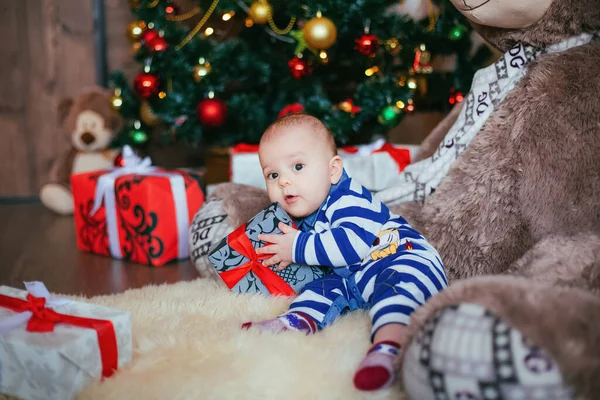 Elinde hediye kutusuyla öfkeli oyuncak ayıya uzanan şirin bir çocuk. Yeni yıl süslemesinde beyaz mavi gömlekli çocuk — Stok fotoğraf