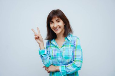 Gülümseyen kadının portresi zafer işareti gösteriyor ve gri bir arka planda izole edilmiş kameraya bakıyor.