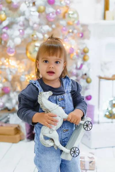 Noel ağacının altında elinde tahta oyuncak atla küçük güzel bir kız çocuğu. — Stok fotoğraf