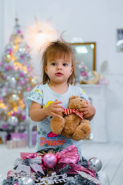Küçük güzel kız elinde oyuncak ayıyla elinde Noel ağacıyla arka planda hediyelerle. — Stok fotoğraf
