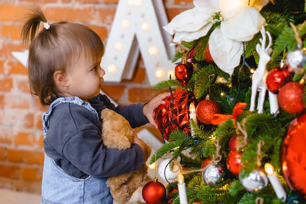 Küçük güzel kız, elinde oyuncak ayıyla Noel ağacını süslüyor. — Stok fotoğraf