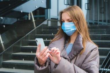 Cerrahi maske takan beyaz bir kadın şehrin dışında yürürken akıllı telefon kullanıyor.